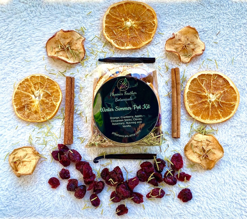 May include: A close-up of a Winter Simmer Pot Kit from Phoenix Feather Botanicals. The kit includes dried orange slices, apple slices, cranberries, cinnamon sticks, cloves, rosemary, nutmeg, and vanilla beans. The kit is packaged in a clear plastic bag with a black label that says "Winter Simmer Pot Kit".