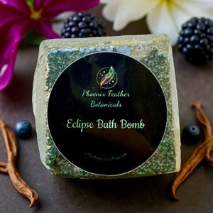May include: A green, square bath bomb with a black circular label reading "Phoenix Feather Botanicals Eclipse Bath Bomb." The image features purple and white flowers, blackberries, and a dark background.