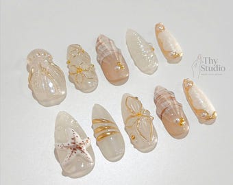 May include: A collection of ten artificial nails with various designs. Some nails feature seashell patterns, gold accents, and small pearls. One nail has a starfish design. The nails are in a variety of shapes and sizes, with a glossy finish. The text "Thy Studio" is visible.