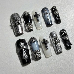 Könnte beinhalten: Ein Set aus zehn Sarg-förmigen Kunstnägeln. Die Nägel weisen verschiedene Designs auf, darunter silberne Kreuze, verzierte Verzierungen und eine Mischung aus Schwarz-, Weiß- und Grautönen. Einige Nägel haben ein glänzendes Finish.