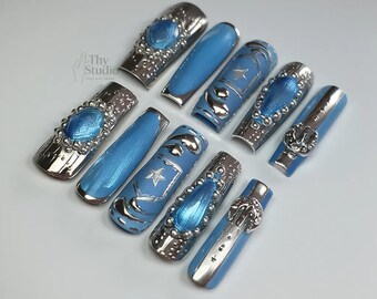Puede incluir: Un conjunto de diez uñas postizas largas en forma de ataúd. Las uñas presentan un color base azul claro con adornos plateados. Algunas uñas tienen una banda plateada con una gema azul, mientras que otras tienen diseños plateados como estrellas y espirales. La marca "Thy Studio" es visible.