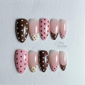 May include: A set of almond-shaped false nails in various designs. Some nails are solid brown with white polka dots, others are pink with brown polka dots, and some have a French tip design with brown or beige accents. The text "Thy Studio" is visible.