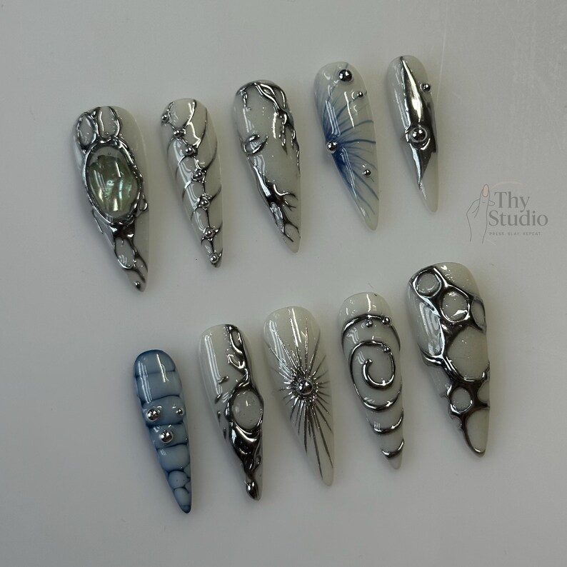 May include: A collection of ten long, almond-shaped artificial nails. Each nail features a white base with silver embellishments, including swirls, dots, and geometric designs. Some nails have blue accents or a central stone.