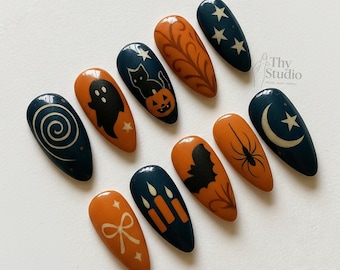 May include: A set of ten almond-shaped Halloween-themed press-on nails. Designs include a ghost, cat with pumpkin, spider, bat, candles, bow, and celestial motifs in orange, navy, and white. The nails are arranged on a white surface.