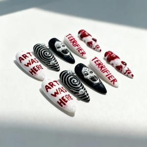Puede incluir: Diez uñas artificiales con varios diseños. Algunas son blancas con el texto "ART WAS HERE" en rojo. Otras son negras con un diseño de cara blanca o espiral. Algunas uñas son blancas con el texto "TERRIFIER" en rojo.