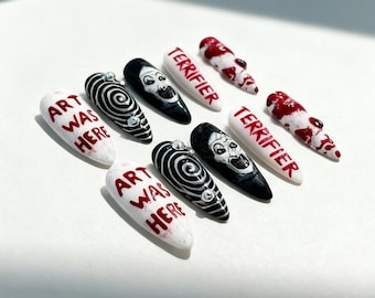 May include: A collection of ten artificial nails with various designs. Some nails are white with the text "ART WAS HERE" in red. Other nails are black with a white face design or a spiral design. Some nails are white with the text "TERRIFIER" in red.