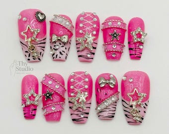 May include: A set of ten pink press-on nails with various designs. Some nails feature star and bow embellishments, while others have a zebra print or a lace-up pattern. The nails are decorated with rhinestones and glitter.