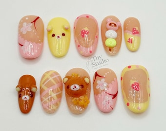 May include: A set of ten artificial nails with various designs. The nails feature a range of colors, including pink, yellow, and orange, with decorative elements such as flowers, bears, and geometric patterns. The text "Thy Studio" is visible.