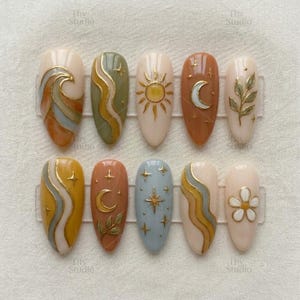Puede incluir: Un conjunto de diez uñas postizas en forma de almendra con varios diseños. Los diseños incluyen olas, soles, lunas, estrellas y motivos florales, todos en una paleta de colores dorados y pastel. Las uñas están dispuestas sobre un expositor de plástico transparente.