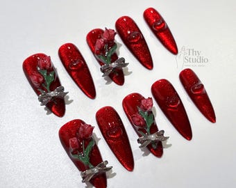 May include: Red glitter nail set with 3D rose and bow accents. Some nails feature a clear heart design. The nails are long, oval-shaped, and arranged on a white surface. The brand name "Thy Studio" is visible.