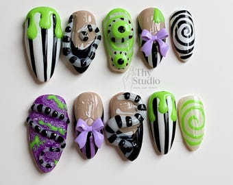 May include: A set of ten press-on nails with a Halloween theme. Designs include black and white stripes, green slime, purple and black snakes, and a black and white spiral. Some nails feature a purple bow.