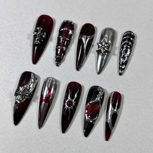 May include: A set of ten long, pointed artificial nails. The nails feature a mix of dark red and silver designs, including crosses, geometric patterns, and metallic accents. The nails have a glossy finish.