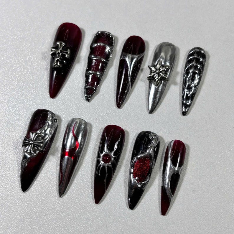 May include: A set of ten long, pointed artificial nails. The nails feature a mix of dark red and silver designs, including crosses, geometric patterns, and metallic accents. The nails have a glossy finish.