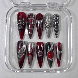 May include: A set of ten long, pointed false nails in a clear plastic case. The nails feature a mix of silver and burgundy designs, including crosses, floral accents, and flame-like patterns. Some nails have a glittery finish.
