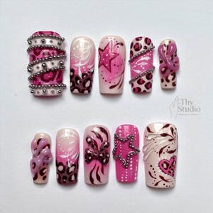 Puede incluir: Un conjunto de diez uñas artificiales con varios diseños. Las uñas presentan una paleta de colores rosa, blanco y marrón. Algunas uñas tienen diseños de estrellas, lazos y estampado de leopardo, mientras que otras están adornadas con cuentas plateadas. El texto "Thy Studio" es visible.