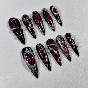 Pode incluir: Um conjunto de dez unhas postiças longas e pontiagudas. Cada unha apresenta uma base vermelha escura com intrincados desenhos prateados, incluindo redemoinhos, pedras preciosas e padrões texturizados. As unhas têm um acabamento brilhante, criando uma estética dramática e gótica.