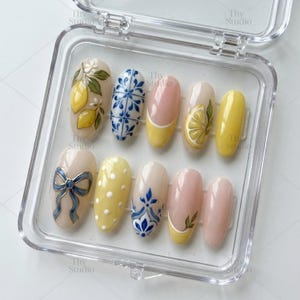 May include: A set of ten oval-shaped press-on nails in a clear plastic case. The designs include yellow lemons with green leaves, blue and white tile patterns, yellow French tips, a blue bow, white polka dots, and solid light pink and yellow colors.