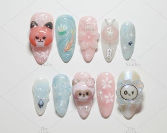 May include: A collection of ten artificial nails with various designs. The nails feature a range of colors, including pink, blue, and white, and are adorned with 3D elements like flowers, animals, and decorative accents. The nails are arranged on a white surface.