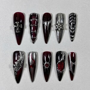 May include: A set of ten long, pointed artificial nails. The nails feature a mix of dark red and silver designs, including crosses, geometric patterns, and a snowflake. Some nails have a gradient effect, transitioning from red to black.