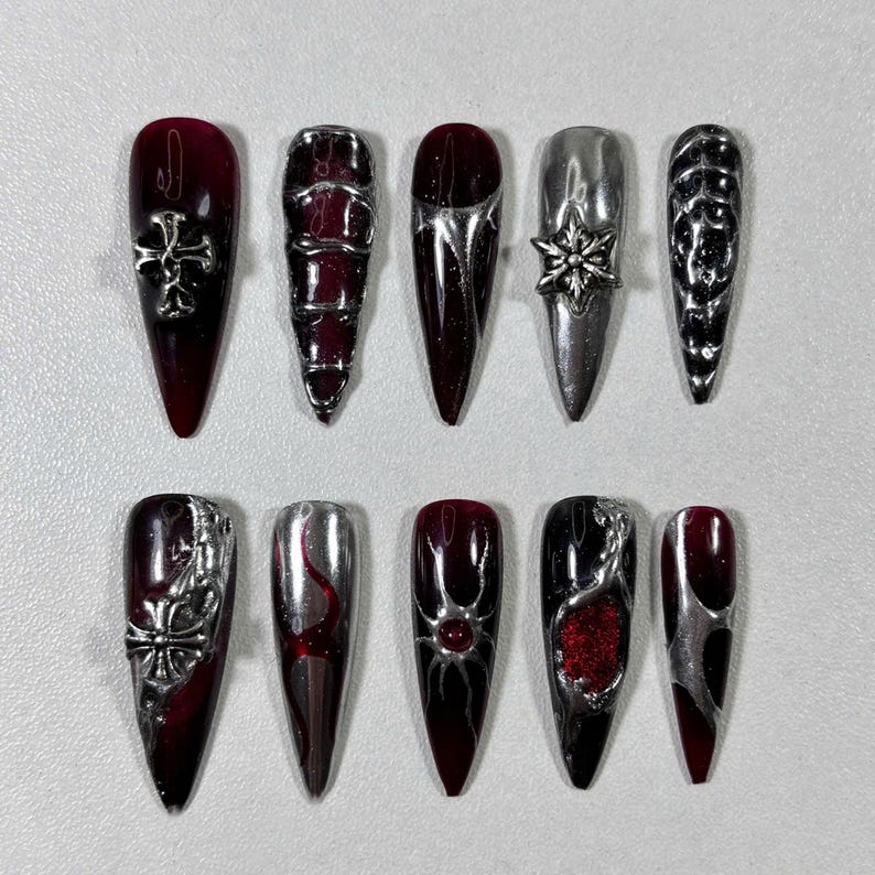 May include: A set of ten long, pointed artificial nails. The nails feature a mix of dark red and silver designs, including crosses, geometric patterns, and a snowflake. Some nails have a gradient effect, transitioning from red to black.