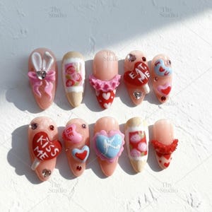 May include: A set of ten artificial nails with various Valentine's Day-themed designs. Each nail features a different embellishment, including hearts, bows, and the phrases "EAT ME" and "KISS ME". The nails are painted in a nude color with decorative elements.