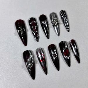 May include: A collection of ten long, pointed artificial nails. The nails feature a combination of dark red and silver chrome finishes. Some nails have silver cross, floral, or geometric embellishments, while others are plain.