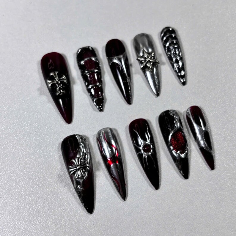 May include: A collection of ten long, pointed artificial nails. The nails feature a combination of dark red and silver chrome finishes. Some nails have silver cross, floral, or geometric embellishments, while others are plain.