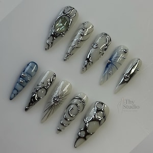May include: A collection of ten long, pointed artificial nails. Each nail features a unique design with silver embellishments, including swirls, dots, and geometric shapes. The color palette includes white, blue, and silver, creating a modern and artistic aesthetic.
