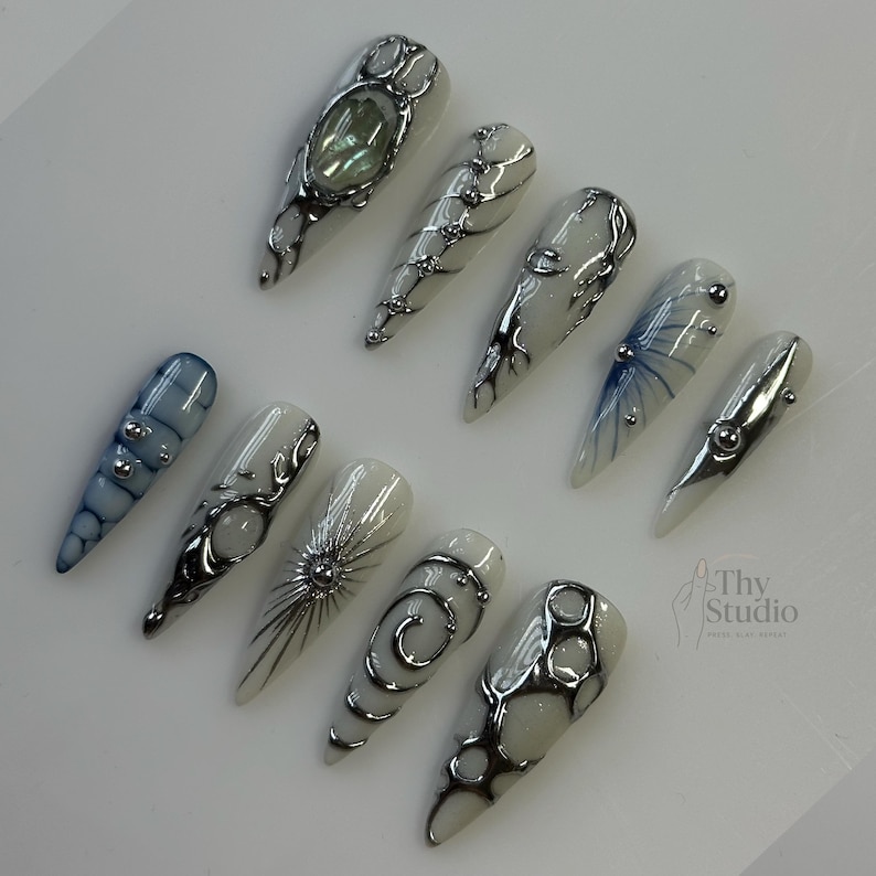 May include: A collection of ten long, pointed artificial nails. Each nail features a unique design with silver embellishments, including swirls, dots, and geometric shapes. The color palette includes white, blue, and silver, creating a modern and artistic aesthetic.