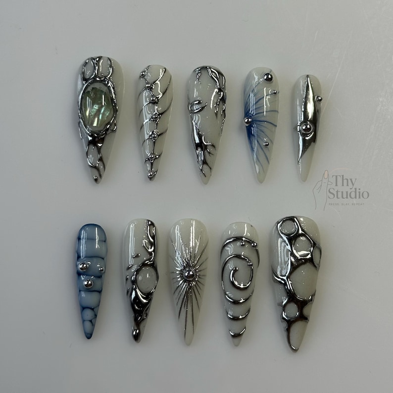 May include: A set of ten long, almond-shaped artificial nails. Each nail features a white base with silver and blue accents. Designs include swirls, lines, and embedded embellishments, creating a unique and artistic look.