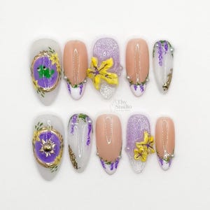 May include: A set of ten artificial nails with various designs. The nails feature a combination of light pink, white, and glittery purple bases. Some nails have floral accents, while others showcase ornate gold and purple embellishments.