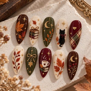 May include: A set of ten almond-shaped press-on nails featuring autumn-themed designs including maple leaves, stacked books, a crescent moon, a black cat, plaid patterns, a coffee cup, and gold star accents in shades of burgundy, cream, and olive green.