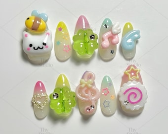 May include: Ten artificial nails with diverse designs. The nails display colours like pink, yellow, and white, and are decorated with 3D embellishments including a bee, cat, musical notes, and stars. The nails are arranged on a white surface.