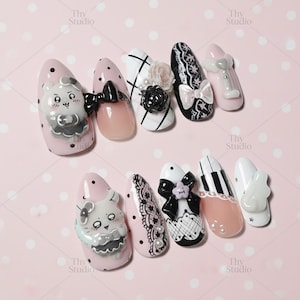 May include: A collection of decorated artificial nails in various designs. The nails feature pink, white, and black colors, with embellishments like bows, flowers, and cartoon characters. Some nails have lace patterns and piano key designs.