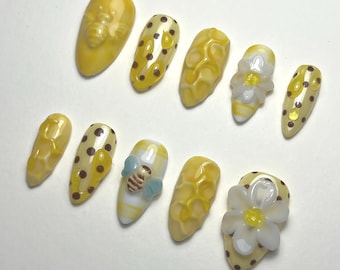 May include: A set of ten yellow and white artificial nails. Some nails feature a bee, polka dots, or a honeycomb design. Other nails have a white flower with a yellow center or a small bee with blue wings.