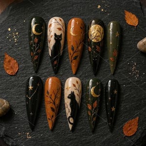 May include: A set of ten almond-shaped press-on nails featuring autumn-themed designs. The nails display black cats, golden crescent moons, full moons, stars, and delicate leaf branches in shades of black, cream, orange, and olive green.