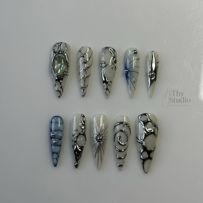 May include: A set of ten long, almond-shaped artificial nails. Each nail features a white base with intricate silver and black designs, including swirls, lines, and geometric patterns. Some nails have blue accents and small silver embellishments. The nails are arranged on a white surface.