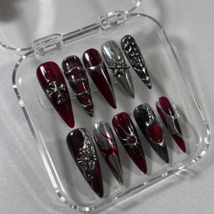 May include: A set of ten long, pointed false nails in a clear plastic case. The nails feature a variety of designs, including silver crosses, metallic accents, and a mix of burgundy, silver, and black colors.