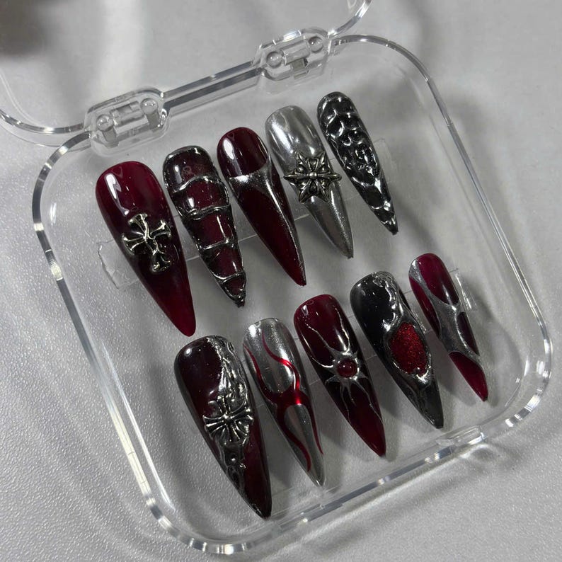 May include: A set of ten long, pointed false nails in a clear plastic case. The nails feature a variety of designs, including silver crosses, metallic accents, and a mix of burgundy, silver, and black colors.