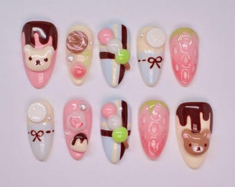 May include: A collection of ten almond-shaped false nails, each decorated with a different dessert-inspired design. The designs feature bears, lollipops, bows, and strawberries, in pink, brown, white, and green. The nails are displayed on a white background.