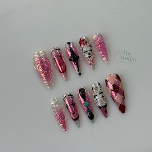 May include: A set of ten long, almond-shaped artificial nails. The nails feature various designs, including a heart, stars, a clown face, and geometric patterns. Colors include pink, red, black, and silver, with iridescent and glitter accents. The nails are arranged on a white surface.