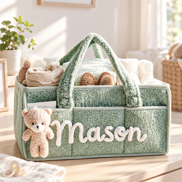 May include: A sage green textured fabric storage caddy with two handles, featuring the name Mason in white script lettering and a small plush teddy bear attached to the front. The caddy contains various baby items including cloths and brushes.