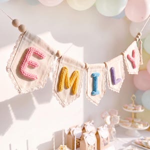 Op de afbeelding: Een decoratieve slinger met vijf off-white stoffen vlaggetjes met kanten rand, geregen aan een koord met houten kralen. Elk vlaggetje toont een verhoogde, grof gebreide letter in roze, geel, blauw, paars en roze, die de naam EMILY spellen.