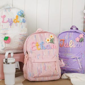 Könnte beinhalten: Drei bunte Rucksäcke mit eingestickten Namen. Der weiße Rucksack hat den Namen "Lydia" in Pastellfarben, einen Erdbeer-Anhänger und blaue Blumenakzente. Der rosa Rucksack hat den Namen "Ethan" und Blumenakzente. Der lila Rucksack hat den Namen "Julia" und Blumenakzente.