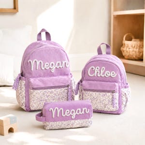 May include: Two lavender corduroy backpacks and a matching pencil case. The larger backpack features the name Megan, the smaller backpack features the name Chloe, and the pencil case features the name Megan. Each item has a floral print base.