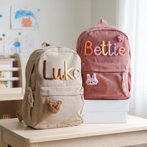 May include: Two corduroy backpacks, one tan and one dusty rose, sit on a table. The tan backpack has the name "Luke" in colorful letters and a teddy bear patch. The rose backpack has the name "Bettie" and a bunny patch.