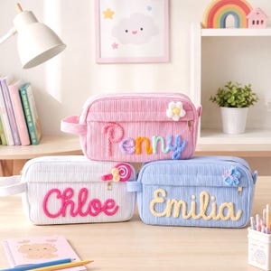 Puede incluir: Tres estuches rectangulares de pana en rosa, blanco y azul claro. El estuche rosa presenta el nombre Penny en letras de lana de colores y un parche de flor. El estuche blanco muestra el nombre Chloe en lana rosa y un parche de piruleta.