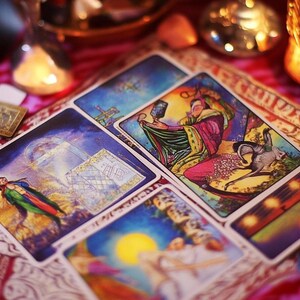 May include: A spread of tarot cards on a patterned fabric with a candle and a glass jar in the background. The cards feature colorful illustrations and intricate designs.