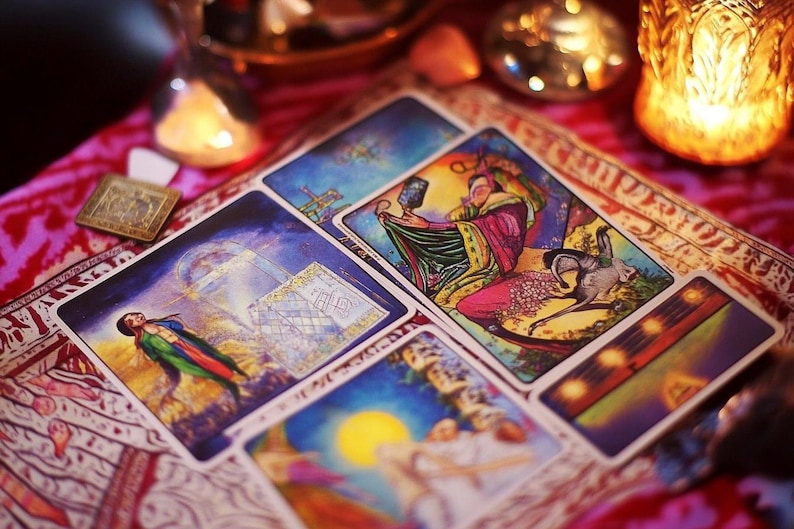 May include: A spread of tarot cards on a patterned fabric with a candle and a glass jar in the background. The cards feature colorful illustrations and intricate designs.