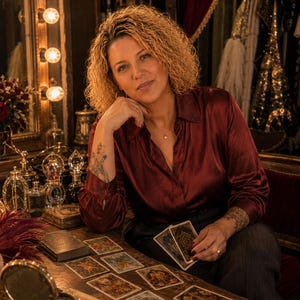 Peut inclure: Une personne aux cheveux blonds boucl&eacute;s portant une chemise en satin rouge fonc&eacute; &agrave; manches longues et boutonn&eacute;e. La personne est assise &agrave; une table en bois avec plusieurs cartes de tarot &eacute;tal&eacute;es, tenant une carte. Un miroir est visible.